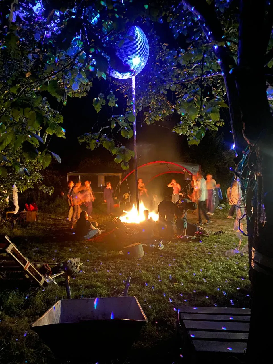 Menschen sitzen und stehen um ein Lagerfeuer. Oben hängt eine Discokugel, die blau angeleuchtet wird.